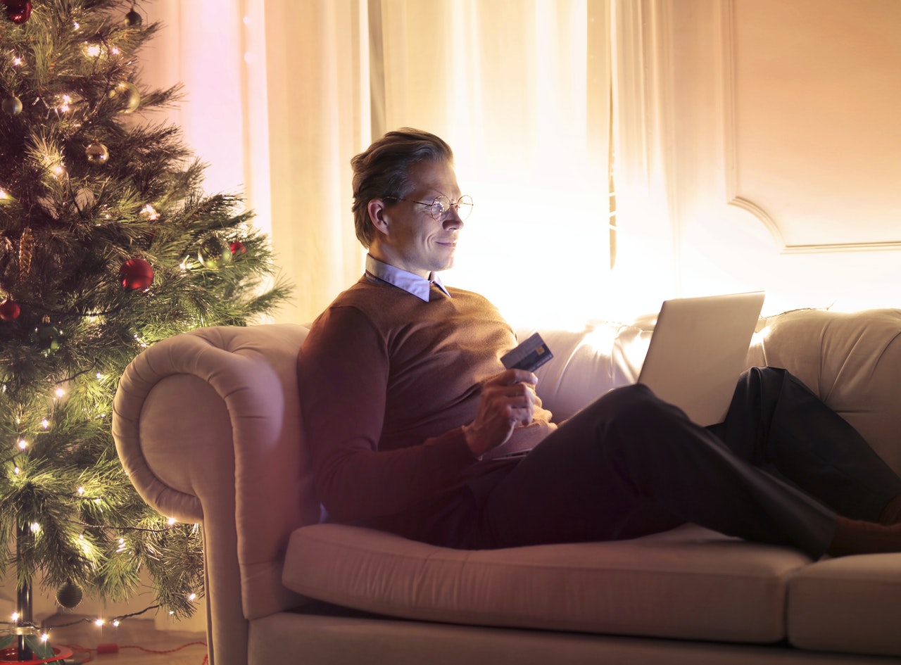Homme d'une cinquantaine d'années assis sur un canapé en train d'acheter en ligne, avec en fond un sapin de Noël